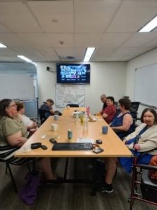 A group of people sitting around a board room table with a screen at the end with more people joining the meeting via zoom.
