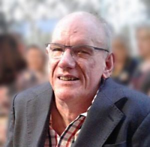 A photo of QDN board member Des Ryan in a dark grey jacket and white shirt with red and blue stripes and wearing glasses and smiling at the camera.