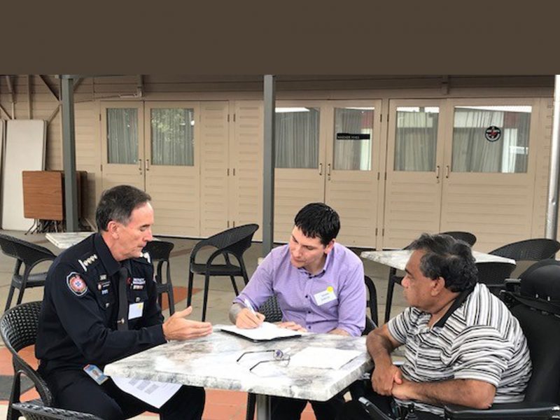 Three men sitting around a table, one in a emergency services uniform. One man taking notes and the man in the uniform is explaining something.