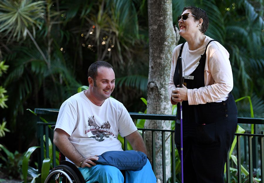 A man in a wheelchair and a woman standing holding a cane are outdoors laughing together.