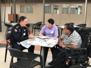 Three men sitting around a table, one in a emergency services uniform. One man taking notes and the man in the uniform is explaining something.