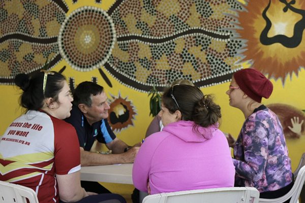 A group of people talking around a table with Aboriginal artwork on the wall behind them.