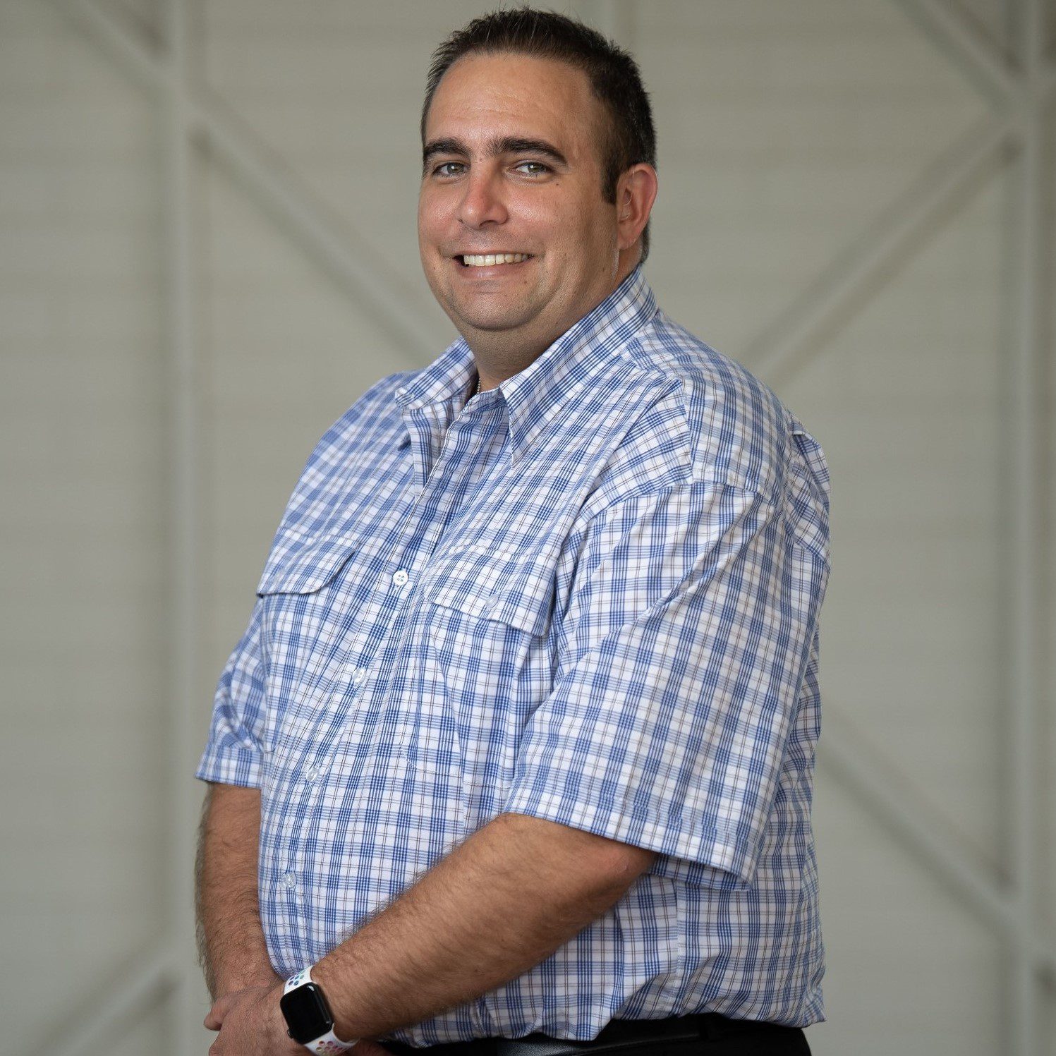 A man with short brown hair, wearing a white and blue checked buttoned up business shirt with short sleeves, he is looking at the camera smiling.