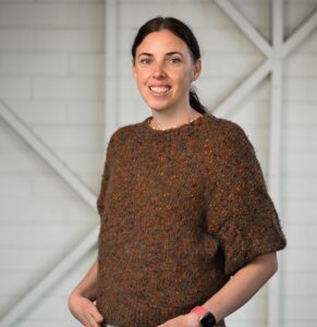 A woman standing in front of the camera with her hands in her front pockets, smiling at the camera. She has dark hair tied back into a low ponytail. She is wearing a brown short-sleeved jumper with light coloured pants.