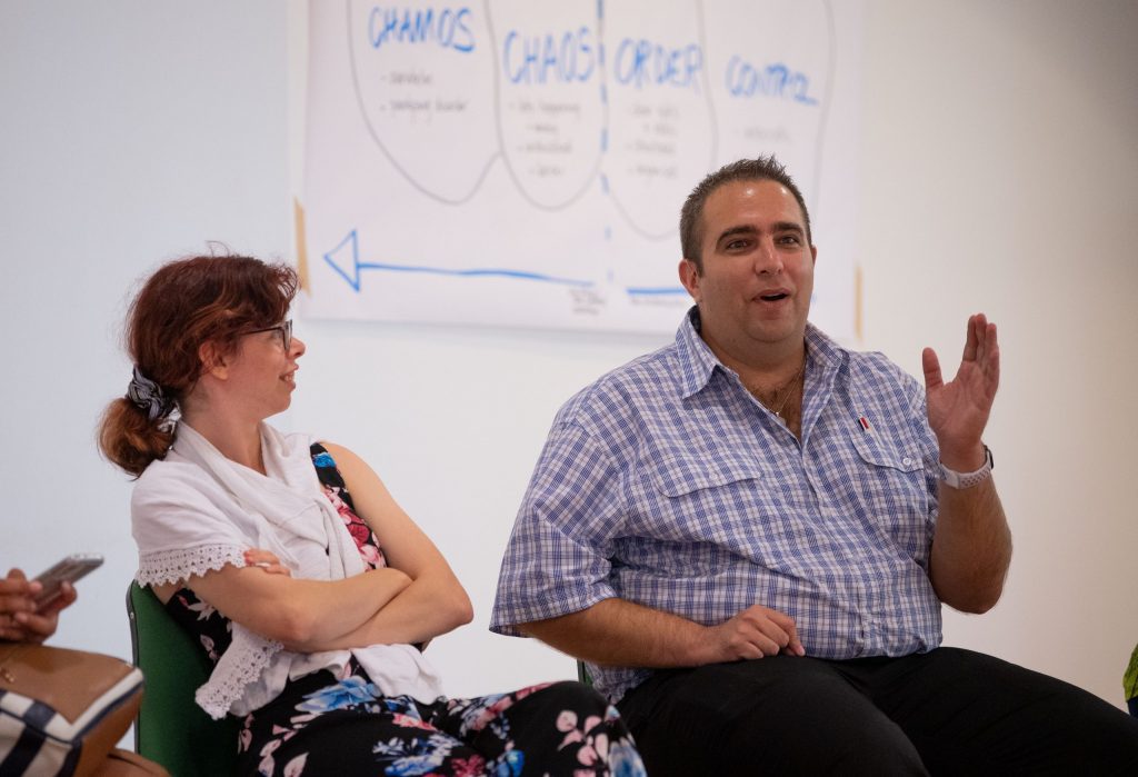 A woman and man sitting next to each other. The man has his hand up and is explaining something to the wider group outside the photo area.