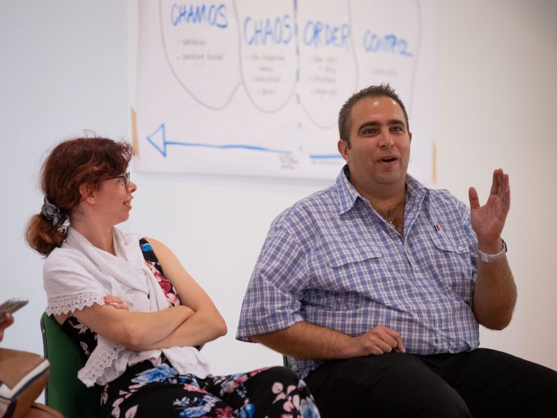 A woman and man sitting next to each other. The man has his hand up and is explaining something to the wider group outside the photo area.