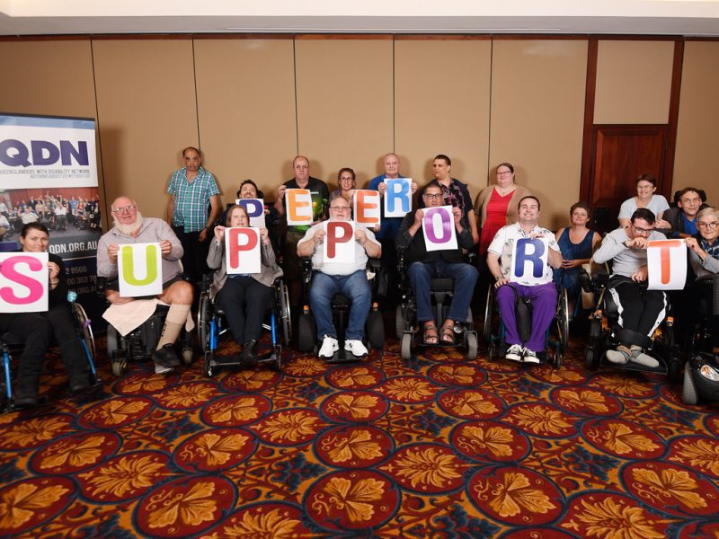 A group of people in wheelchairs and a group of people standing behind them are holding up colourful letters that spell Peer Support.