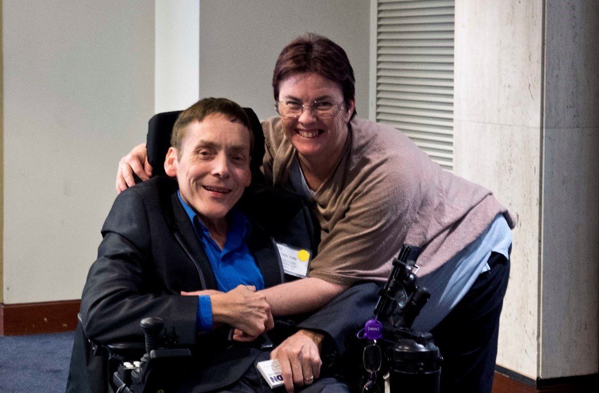 A man in a wheelchair is being embraced by a lady standing next to him. They are both smiling.