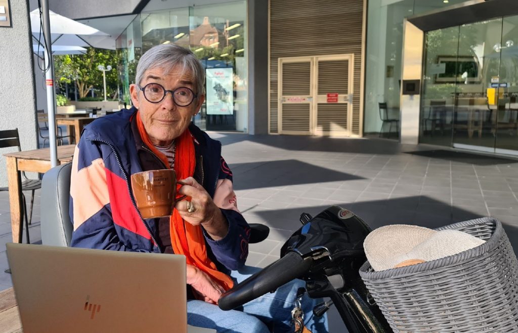 A lady sitting on an electric scooter is holding a cup of coffee with a laptop on the table in front of her.