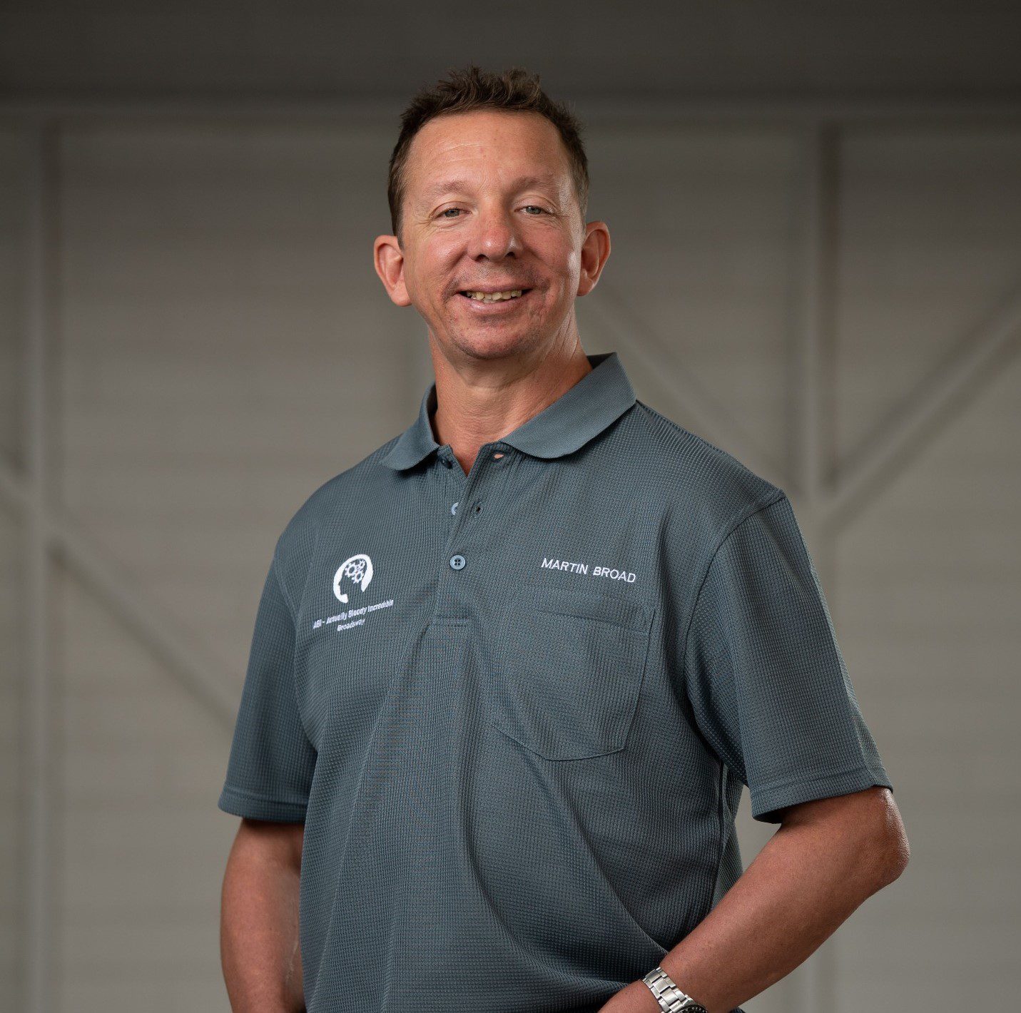 A man standing with his hands in his pant pockets, wearing a grey polo and smiling at the camera.