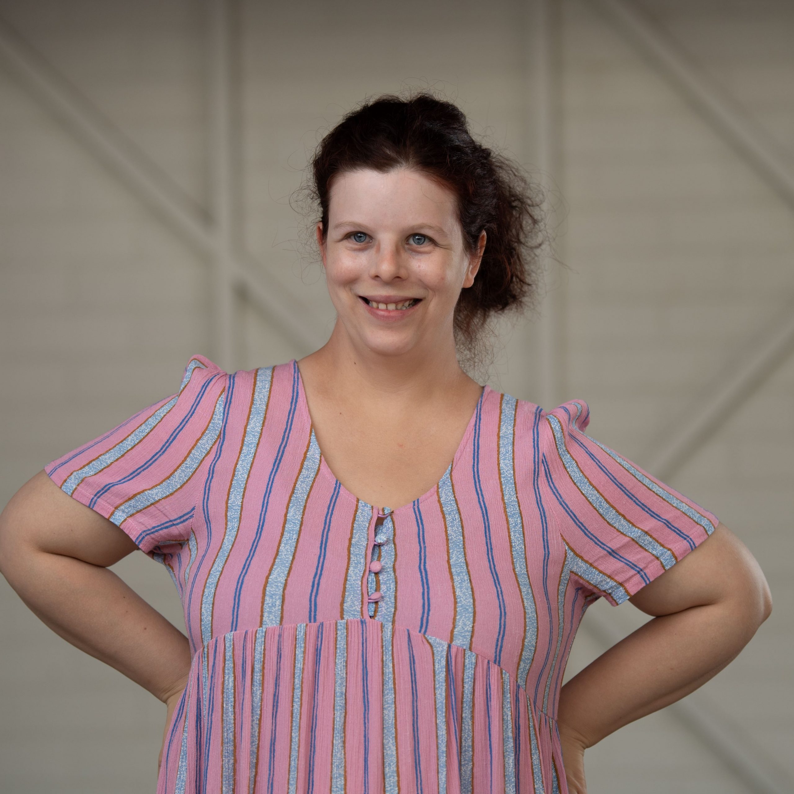 A woman with dark brown curly hair pulled back wearing a pink and blue stripped dress, with her hands on her hips smiling at the camera.