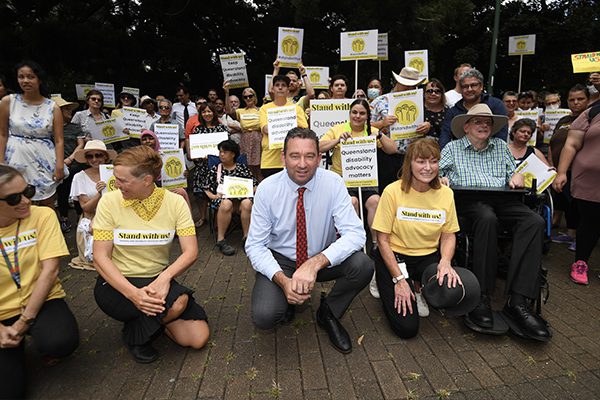 A group of people outdoors, holding placards and wearing yellow tops that say Stand with us! Queensland disability advocacy matters.