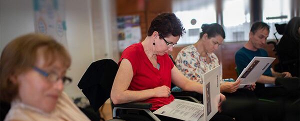 Three women in wheelchairs and a man in a chair are reading information in some white folders. You cannot see the content in the folders.