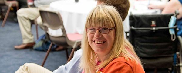 A women with shoulder length blonde hair, glasses and a red shirt is smiling widely at the camera. She is sitting at a table. In the background are blurred tables with groups of people sitting around them.