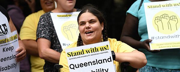 A girl with a yellow shirt is holding a Stand with us campaign poster with people behind her at a rally.