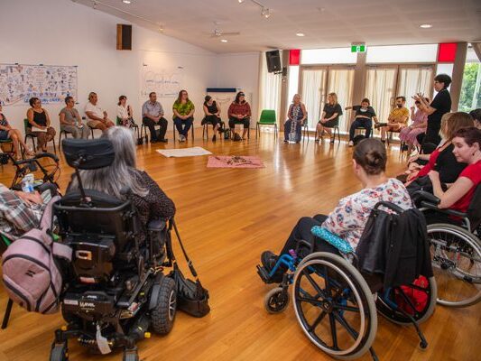 A group of people are gathered in a cirecle. They are all seated down facing the middle as a single lady is standing and presenting to the group. Some of the people are in wheelchairs.