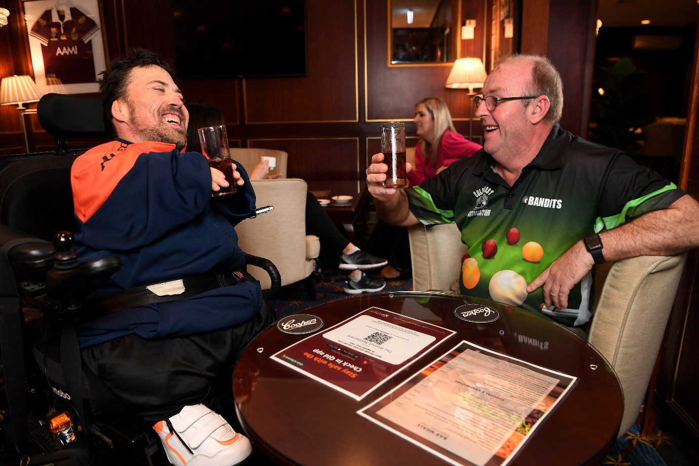 Two men having a drink. The man on the left is in a wheel chair and wearing a blue and orange shirt and the man on the right is wearing a black and green branded shirt. They are in a bar lunge with a table in front of them and are both laughing.