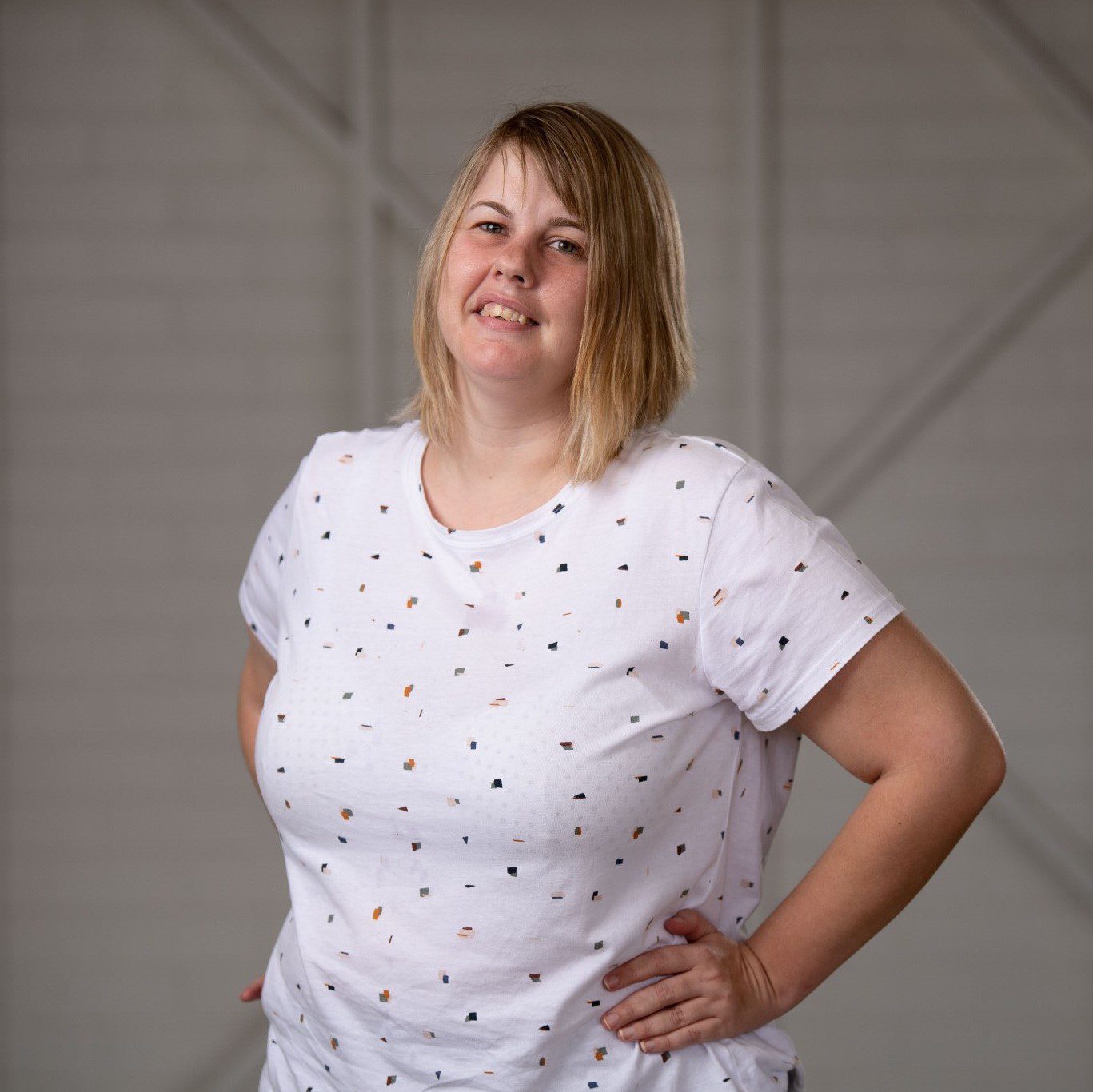 A woman in a white tshirt with small pattern on it, with dark blonde hair, standing with her hands on her hips smiling at the camera.