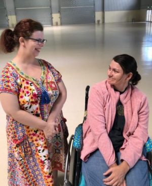 A lady in a wheelchair is smiling to a lady standing next to her. They are in a large hall.