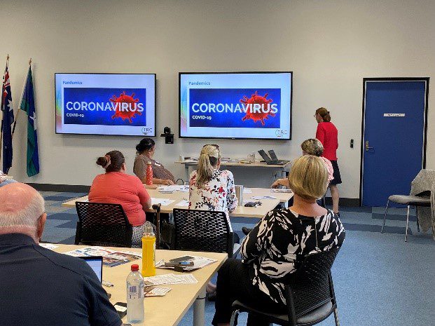 The backs of a group of people sitting at tables looking at screens on the front wall of the room. There is a presenter wearing a red jacket and the screens have Corona Virus written on them.