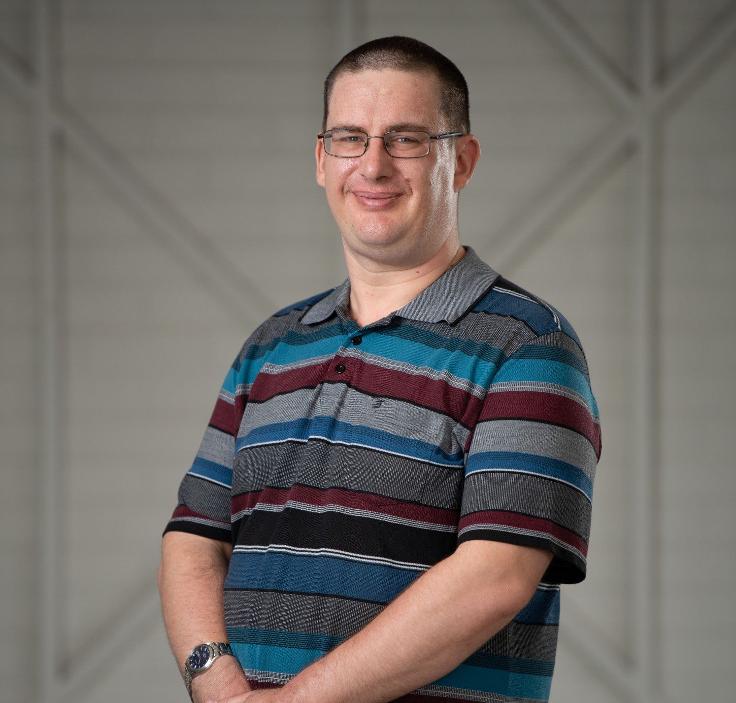 A man standing alone in front of a white wooden wall. He is wearing a Grey, maroon and blue striped shirt and glasses. He is smiling at the camera.