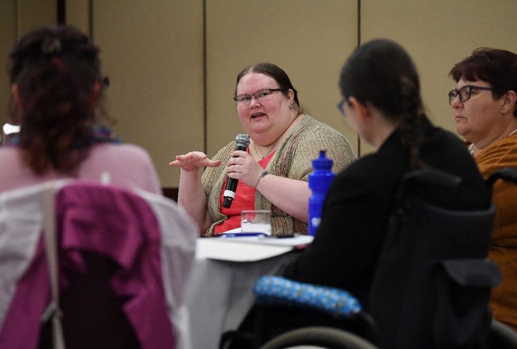The back of two women is out of focus. In focus in a lady sitting at a table talking into a microphone to a group.