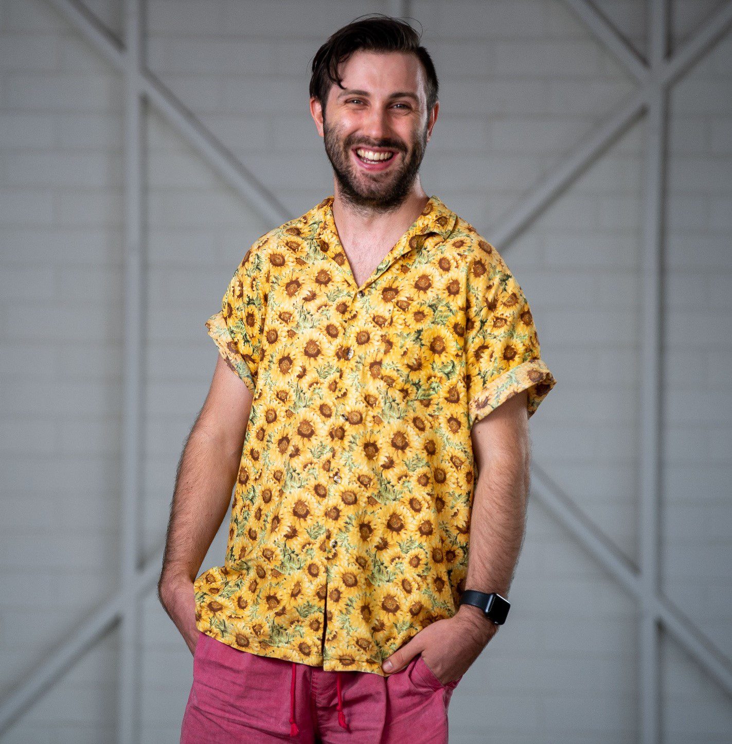 A man standing alone in front of a white wooden wall. He is wearing a bright yellow button up shirt with patterns and red pants. He is smiling broadly at the camera.