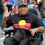 A man in a wheelchair is wearing a black shirt with the Aboriginal flag on the front, wearing a black hat and glasses waving his hand and smiling at the camera.