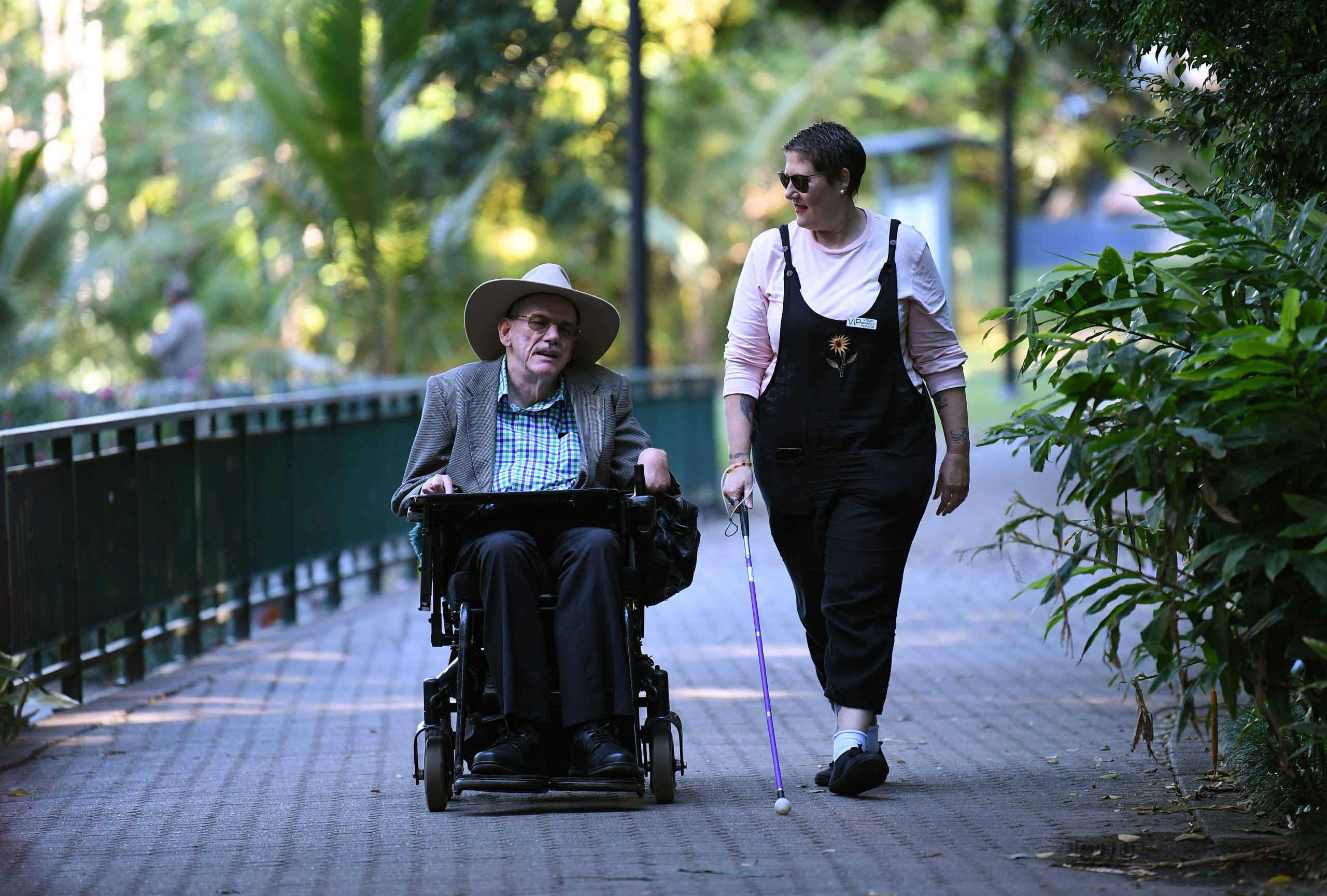 Two people chatting outdoors, walking down a path lined with trees. One in a wheelchair, the other with a cane.