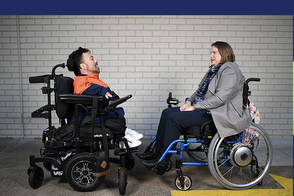 A man and a woman both in wheelchairs, facing each other talking in front of a white brick wall.