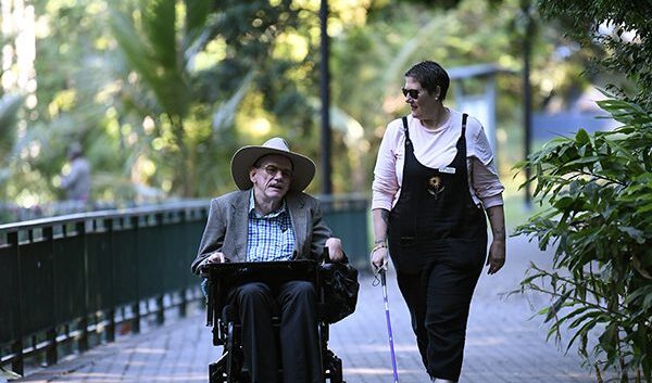 Two people chatting outdoors, walking down a path lined with trees. One in a wheelchair, the other with a cane.