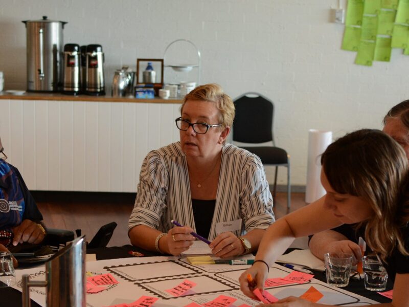 A man in a wheelchair and a woman are sitting around a table. On the table is a white piece of paper covered in pink sticky notes with words. Another lady is leaning over the table to put a note on the paper.