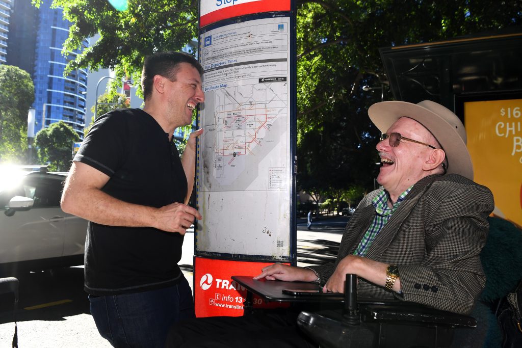 An elderly man in a wheelchair is laughing with a young man who is standing up. They are standing next to a bus stop map in Brisbane City.