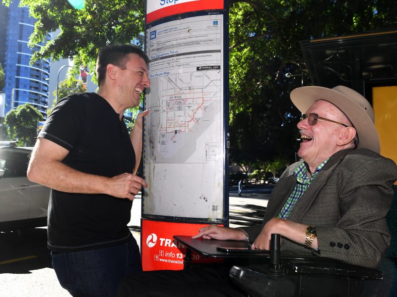 An elderly man in a wheelchair is laughing with a young man who is standing up. They are standing next to a bus stop map in Brisbane City.