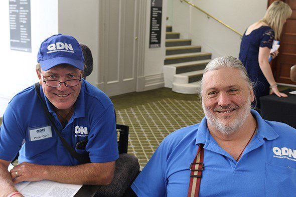 Two men sitting next to each other. Both wearing light blue QDN polos and the man on the left is wearing a matching QDN hat.