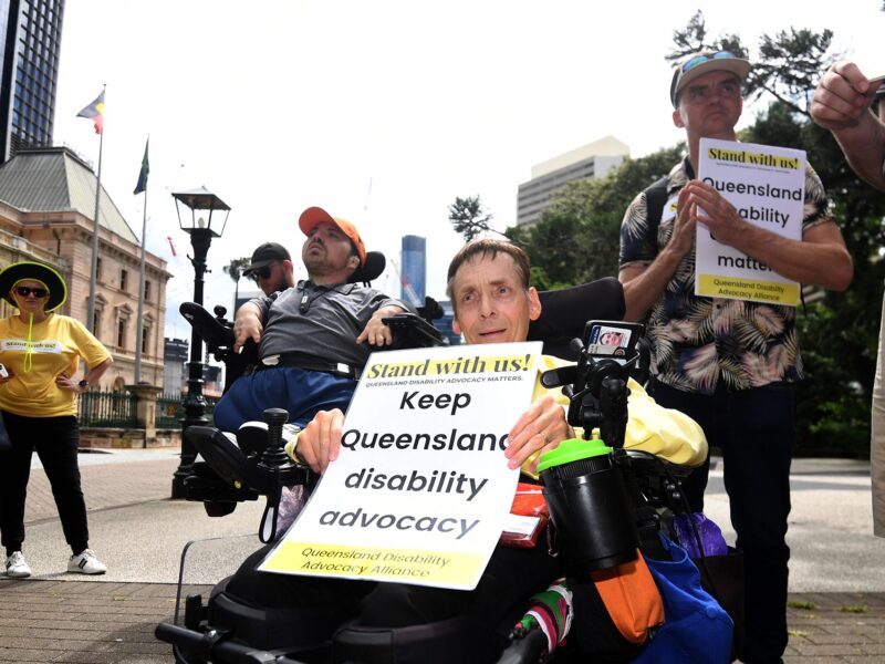 Outdoors in a town square, an older man in a wheelchair is holding a sign saying Keep Queensland disability advocacy. Another man in a wheelchair is behind him and there are other people in the background.