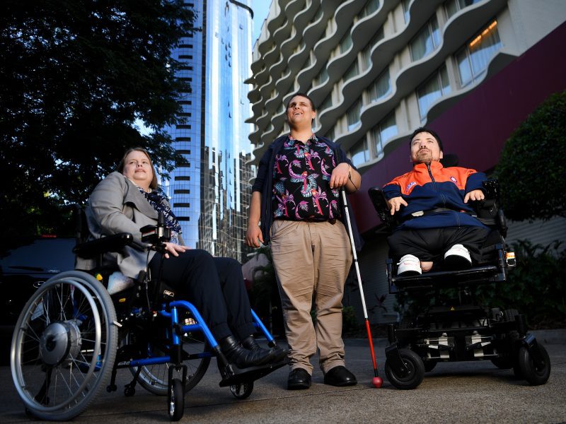 A lady in a wheel chair next to a man holding a white cane and another man in a wheel chair next to him. They are all looking at something behind the camera.
