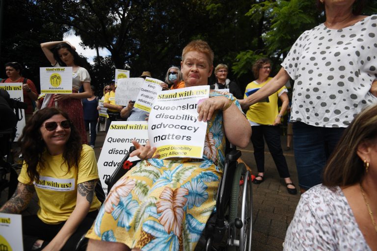 A lady in a wheelchair at the Stand with Us Rally! She is holding a sign with the words, Queensland disability advocacy matters. A lady standing next to her has her hand rested on the back of the wheelchair. They are surrounded by other protesters.