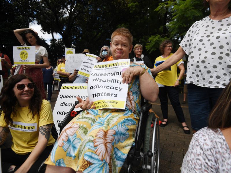A lady in a wheelchair at the Stand with Us Rally! She is holding a sign with the words, Queensland disability advocacy matters. A lady standing next to her has her hand rested on the back of the wheelchair. They are surrounded by other protesters.