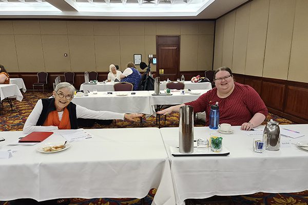 two women sitting at a table at a workshop with their arms reaching towards each other to show they are social distancing,