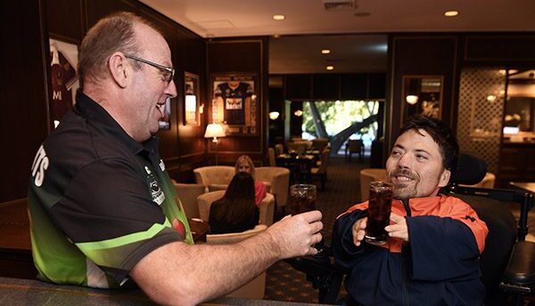 Two men having a drink in a lounge bar. The man on the right is in a wheelchair looking up and smiling towards a man standing up. They are holding their drinks out to one another.