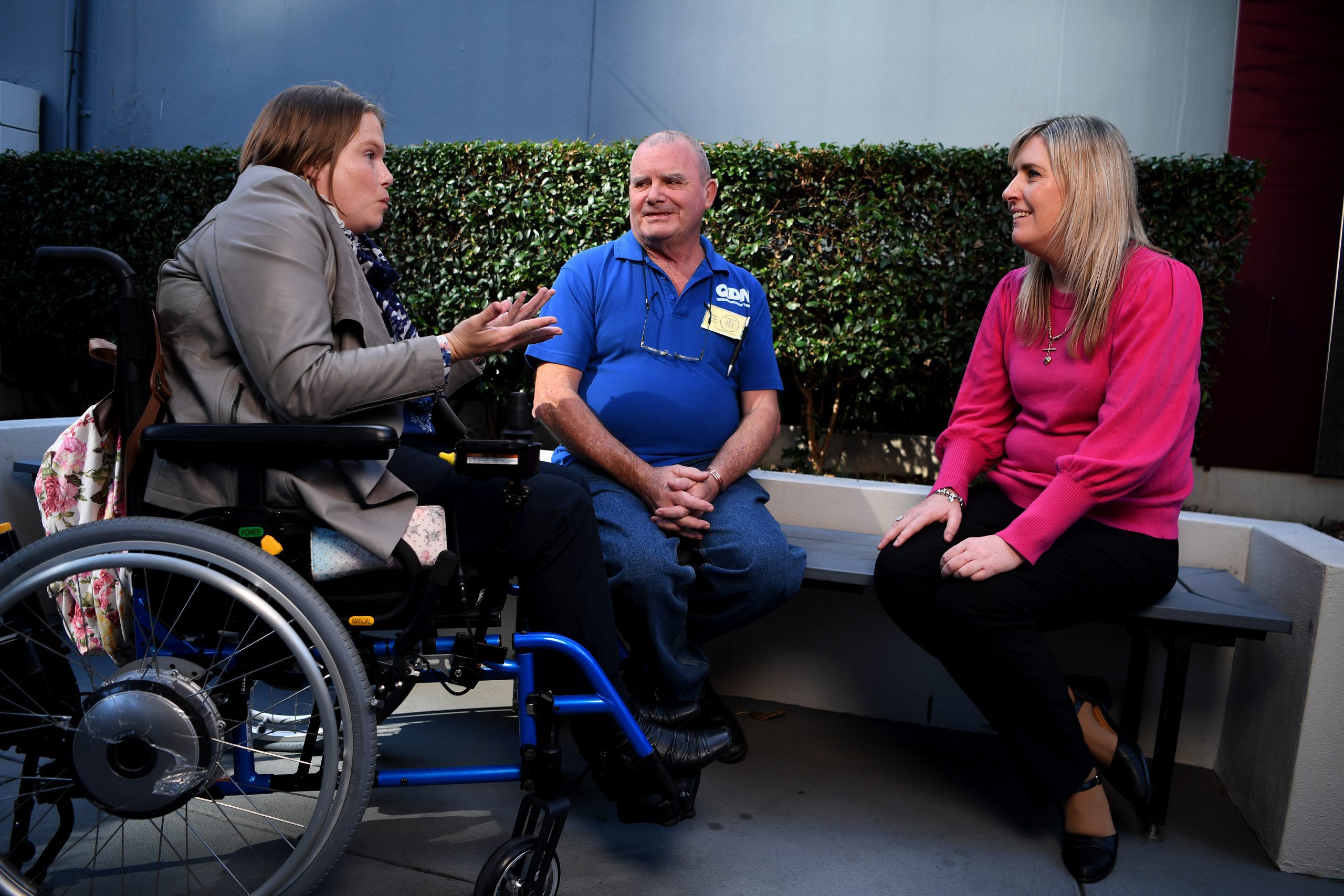 A woman in a wheelchair is holding her arms out in expression, and talking towards another man and woman who are seated on a bench in front of a bush and are looking at her.