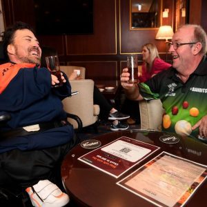 Two men having a drink. The man on the left is in a wheel chair and wearing a blue and orange shirt and the man on the right is wearing a black and green branded shirt. They are in a bar lunge with a table in front of them and are both laughing.