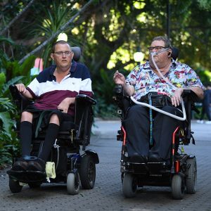 Two men talking while going down a path surrounded by gardens. Both men are in wheelchairs.