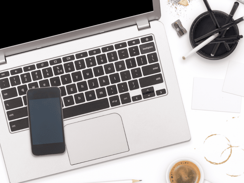 A photo of a laptop with a phone sitting on the keyboard, there are pencils in a holder and a coffee mug with coffee in it beside the laptop.