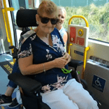 A woman in a wheelchair wearing sunglasses and smiling at the camera, on a bus with another woman behind her popping her head around smiling.
