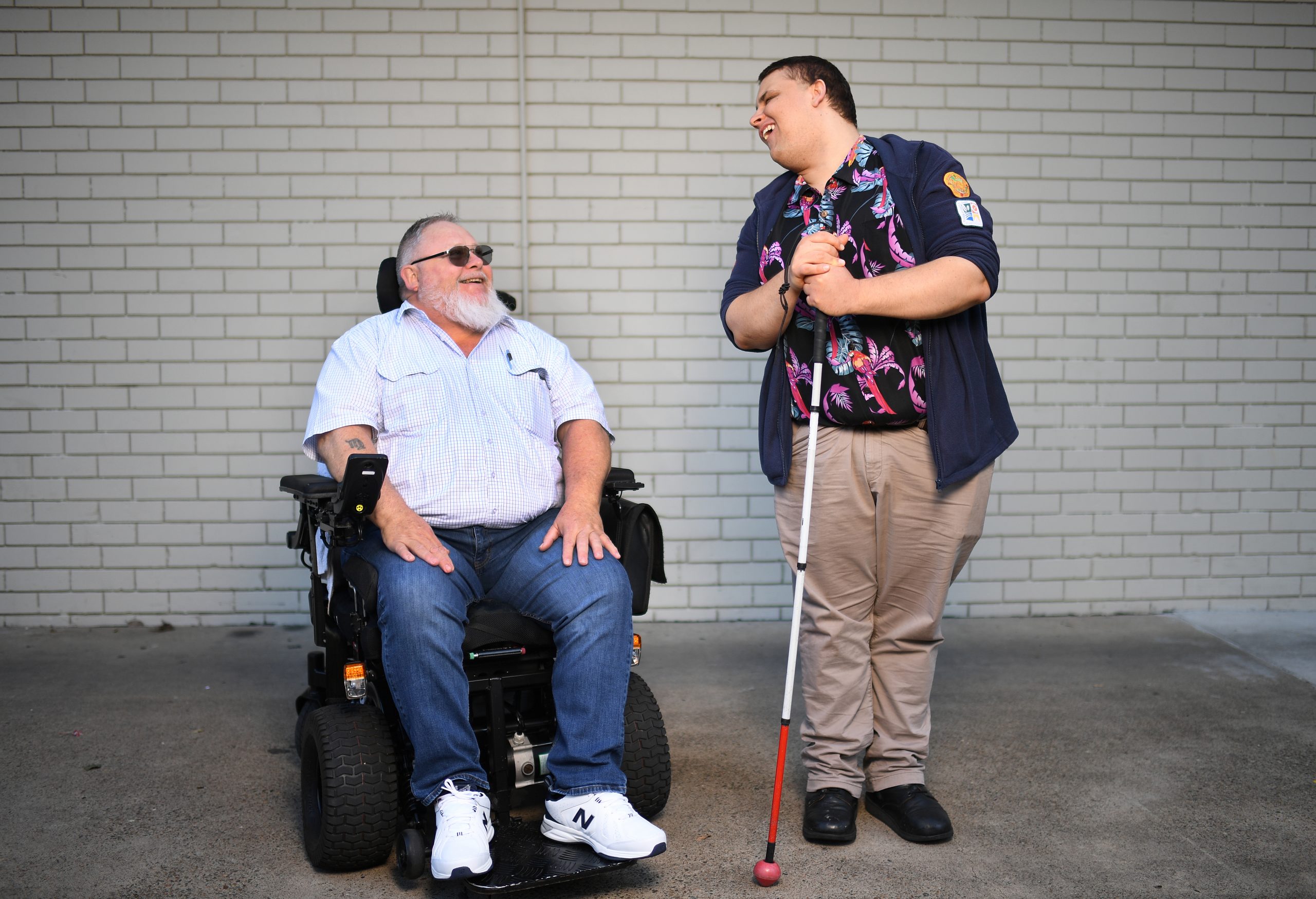 A man in a wheelchair is looking up and smiling at a man standing beside him, who is smiling and looking down. They are in front of a white brick wall.