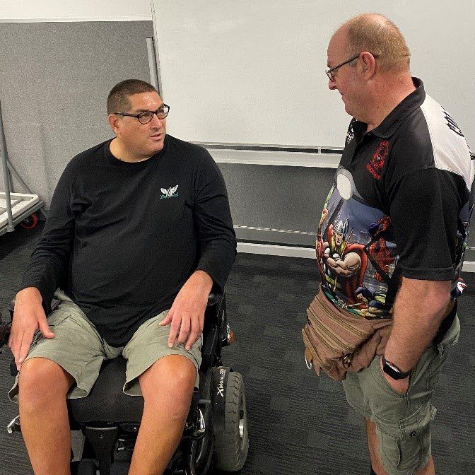 A man in a long sleeved black shirt, shaved dark hair and glasses, sitting in a wheel chair is talking to a man standing next to him who is wearing shorts and a black and white polo.