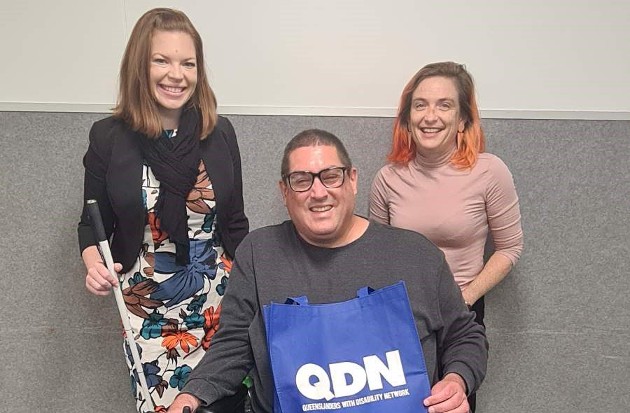 Two women standing and one man sitting in front of them. They are all smiling at the camera. The man has a QDN bag in his lap.
