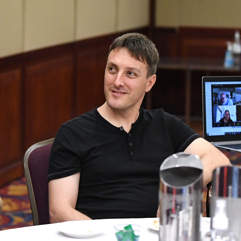 A young man smiling wearing a black shirt is seated at a table listening at a conference.